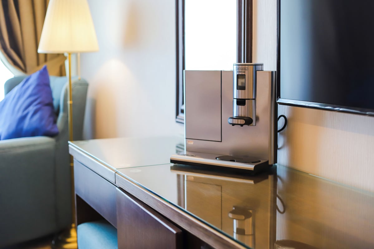 A modern hotel room with a sleek, metallic minibar, a glass-top desk, and a comfortable seating area with a sofa and a lamp.