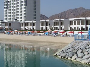 A beachfront hotel with multiple-story buildings, a sandy beach with sun loungers and umbrellas, and a rocky shoreline. - Mirage Bab Al Bahr Hotel (Dhadnah)