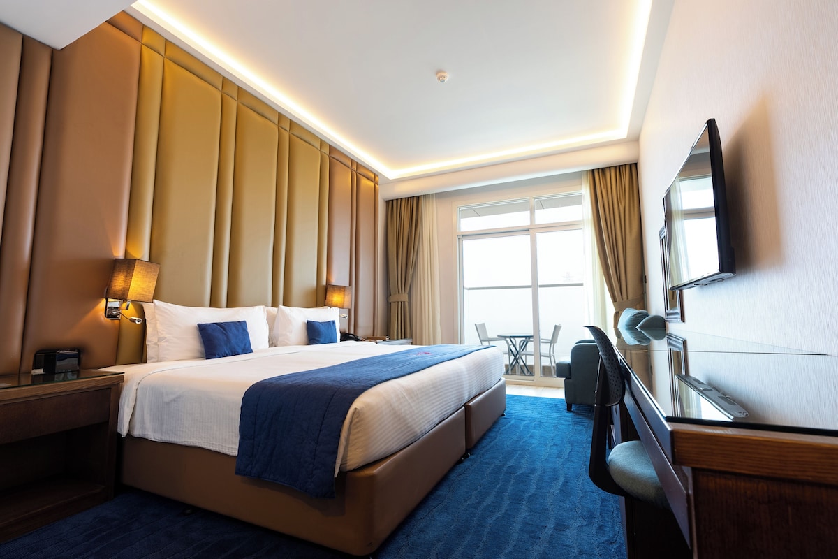 A hotel room with a large bed, a desk, a chair, a TV, and a balcony with a view.