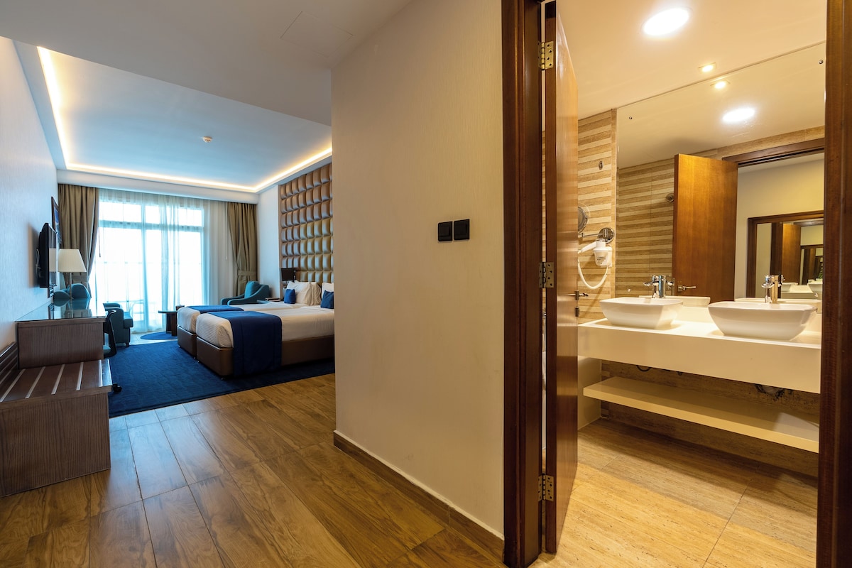 A hotel room with a bed, a desk, a chair, a sofa, a bathroom with a sink and mirror, and a wooden floor.