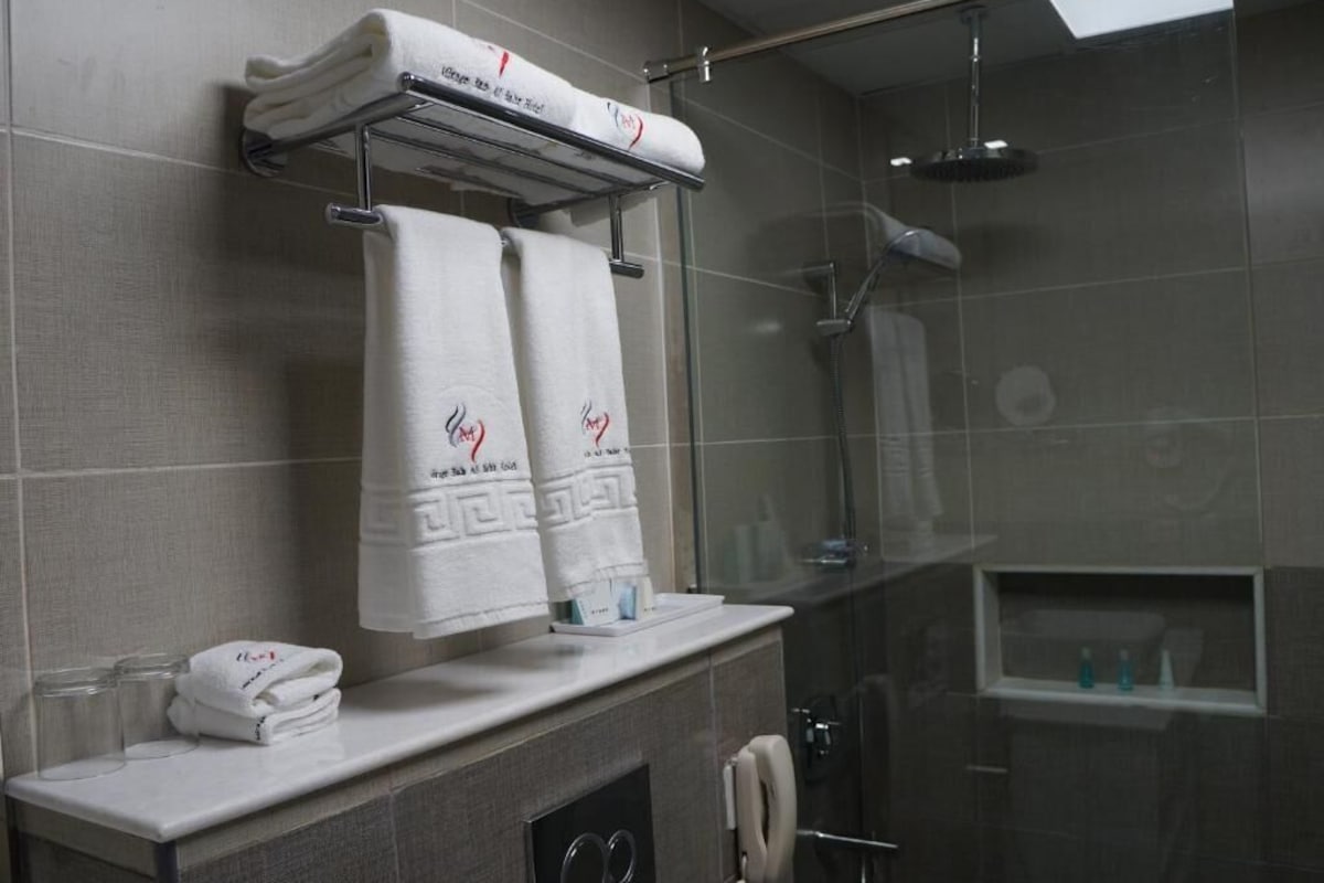 A bathroom with a glass shower enclosure, a towel rack with folded towels, a telephone, and a small shelf with toiletries.