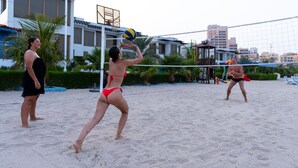 A beach volleyball game with players in red and black bikinis, a volleyball net, and palm trees in the background. - Mirage Bab Al Bahr Hotel (Dhadnah)