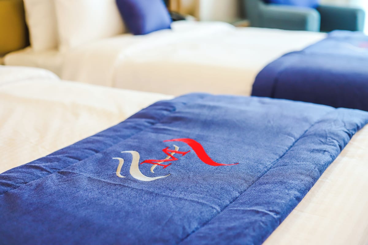Ein Hotelzimmer mit einem Bett, ausgestattet mit einer blauen Bettdecke mit rotem und weißem Logo, einem weißen Kopfteil und einem blauen Kissen.