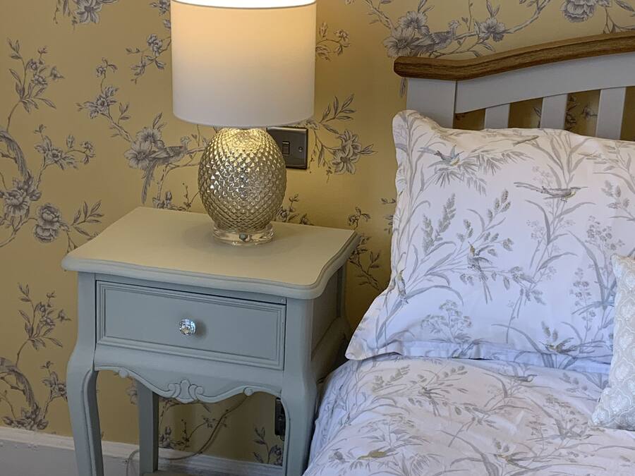 A bedroom with floral wallpaper, a bedside table with a lamp, and a bed with patterned bedding.