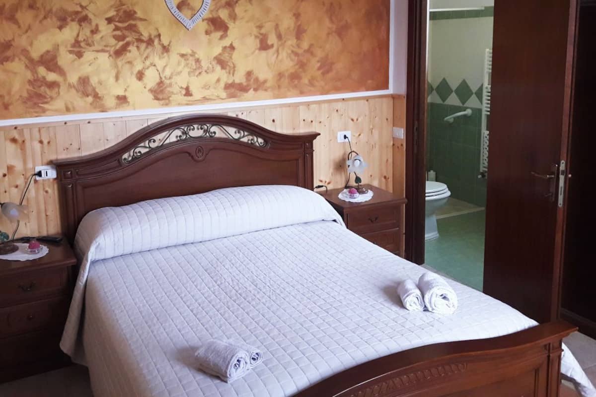 A neatly made bed with a quilted white duvet, two bedside tables with lamps, a wooden headboard, and a wooden wardrobe.