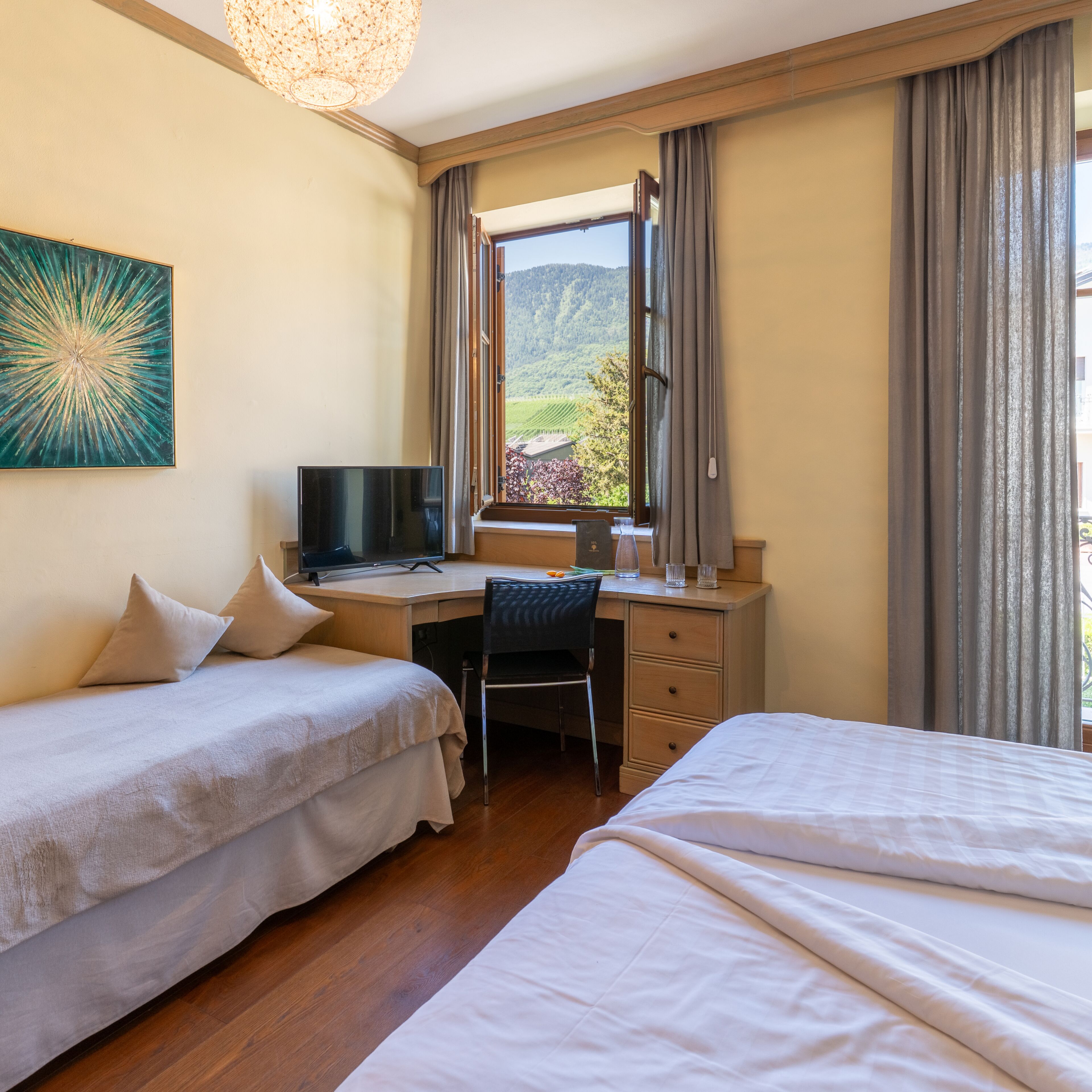 Ein Hotelzimmer mit zwei Betten, einem Schreibtisch mit Computer, einem Sessel, einem Fernseher und einem Fenster mit Vorhängen.