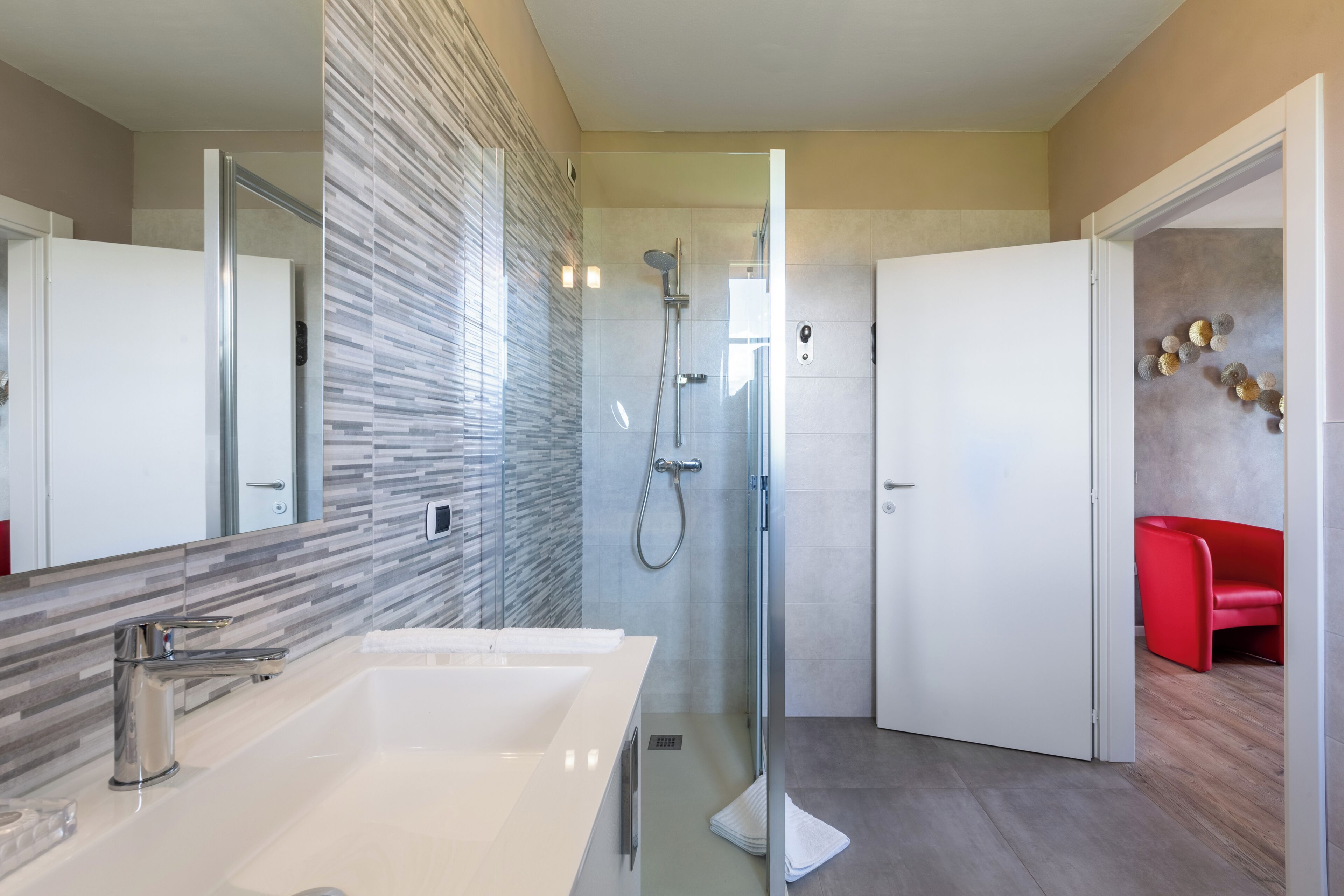 A modern bathroom with a glass-enclosed shower, a double sink vanity, and a red chair in the corner.