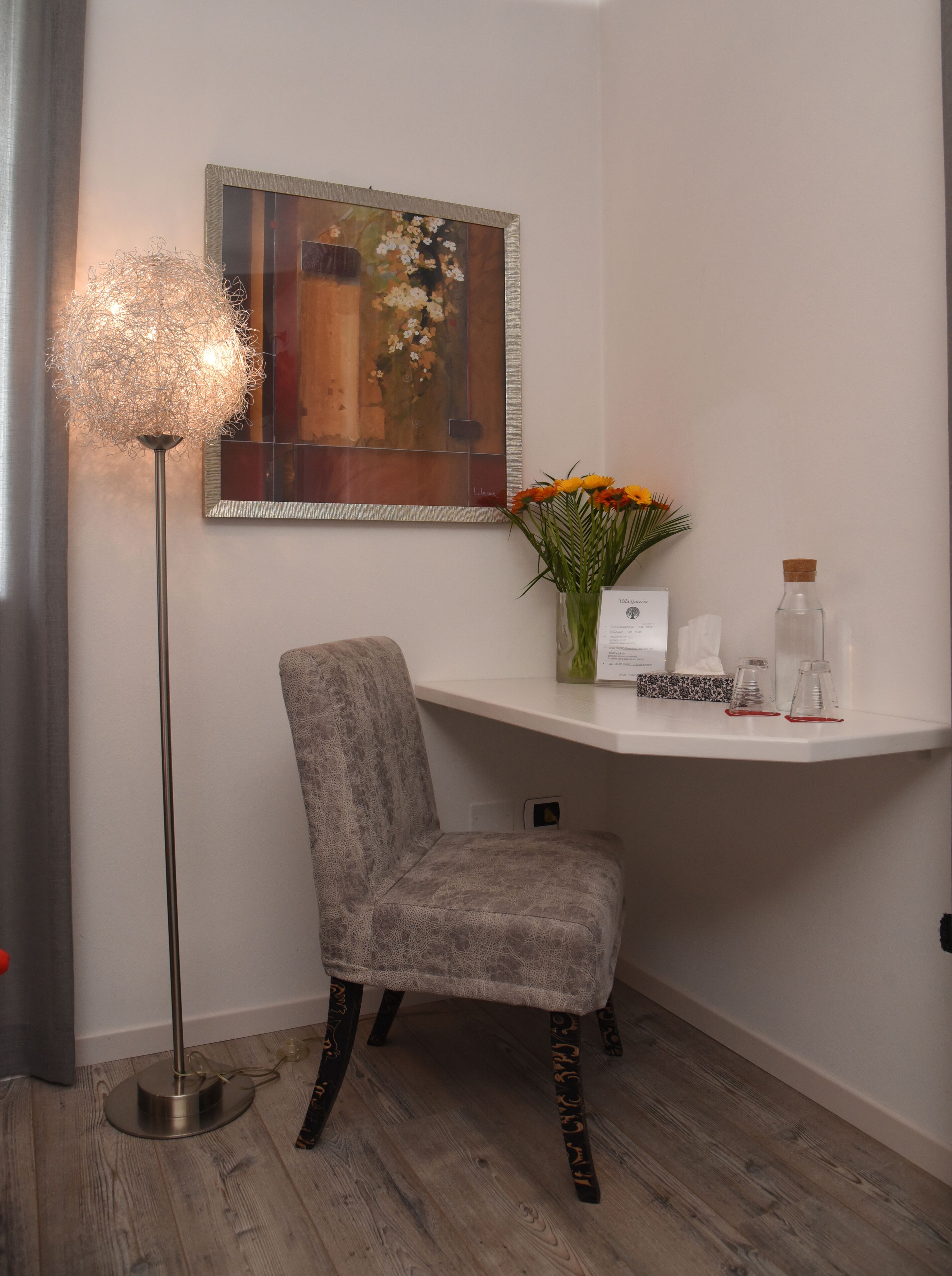 Habitación de esquina con una silla tapizada de gris, una lámpara de pie, un cuadro, un jarrón con flores y un estante pequeño con objetos.