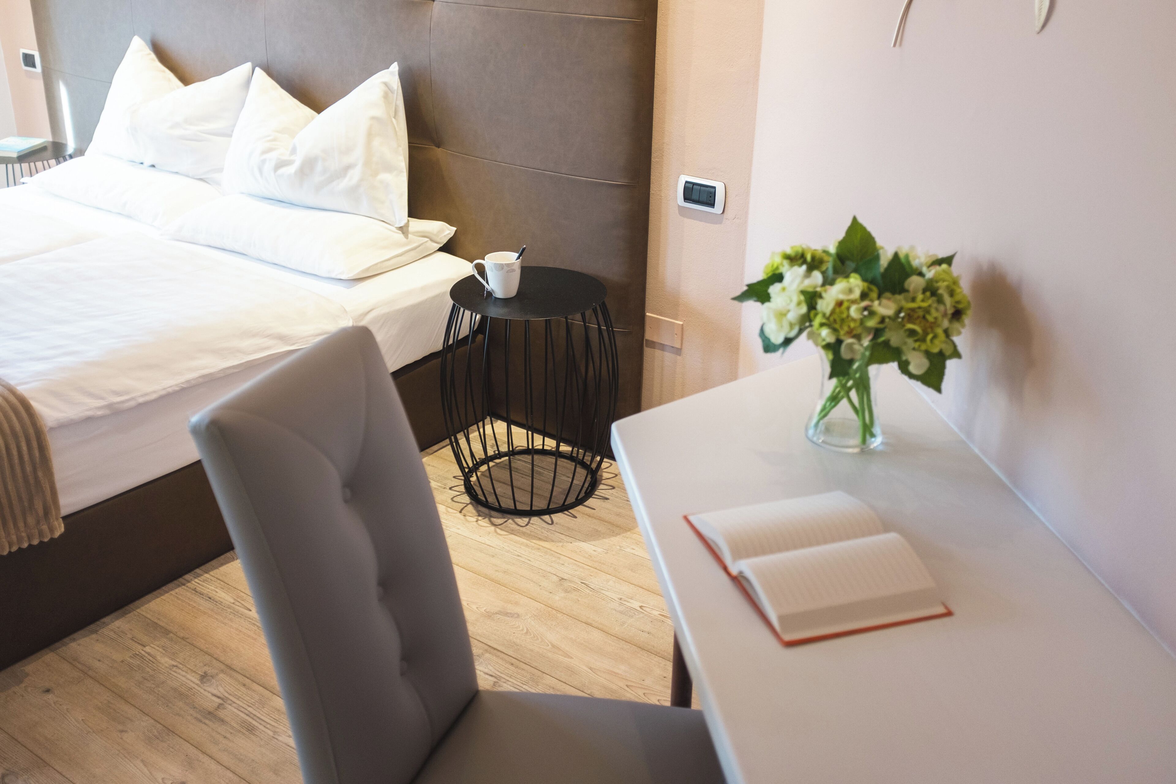 A hotel room with a bed, a bedside table with a book, a chair, a small table with a cup, and a vase with flowers.