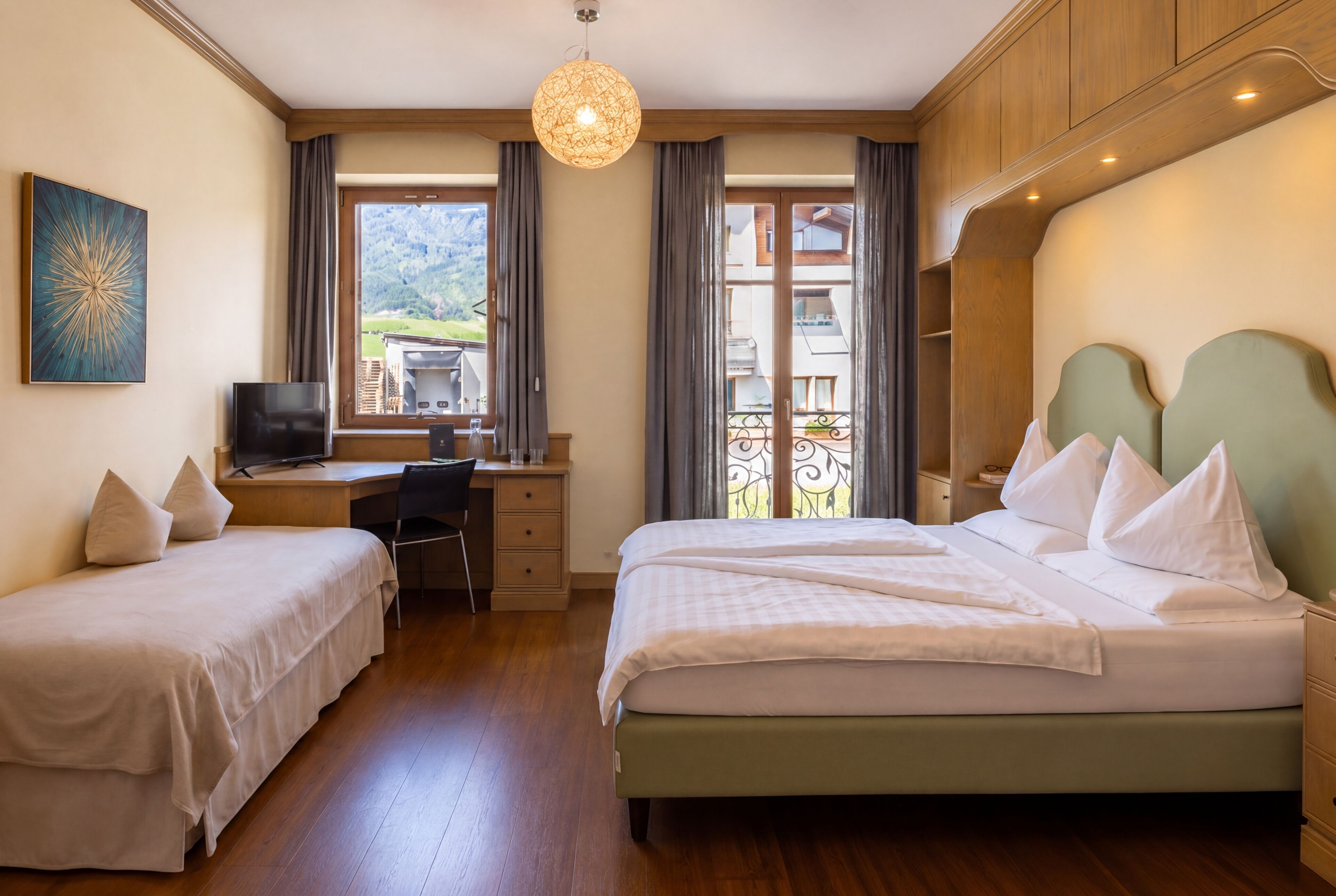 A hotel room with a bed, a desk, a chair, a TV, and a balcony with a view of the mountains.