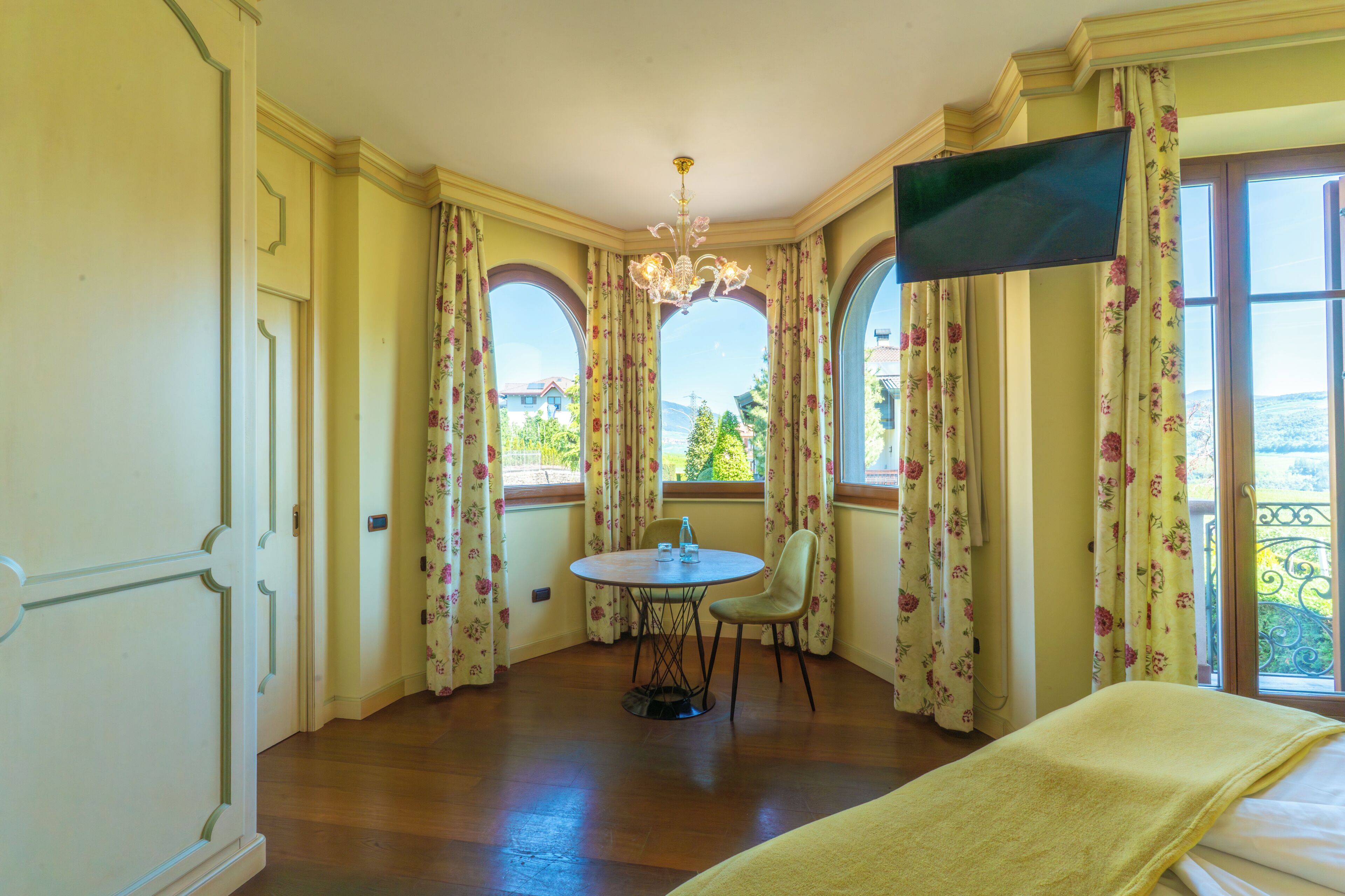 A room with a bed, a round table with chairs, a chandelier, and large windows with floral curtains.