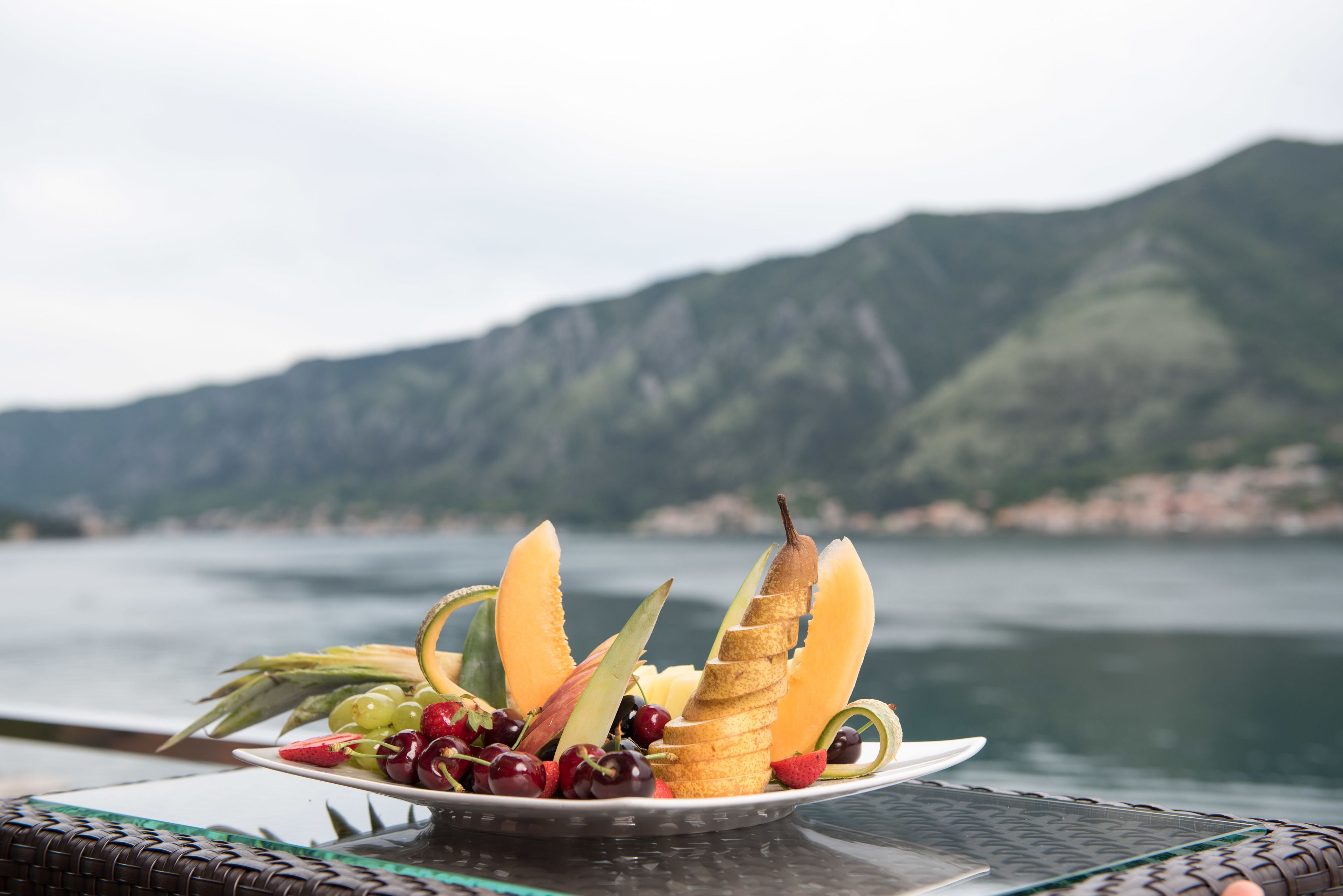 A plate of fruit and bread on a table with a view of a lake and mountains in the background.