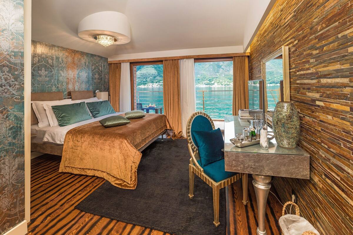 A hotel room with a bed, a desk, a chair, a large window with a view of the sea, and a wooden wall.