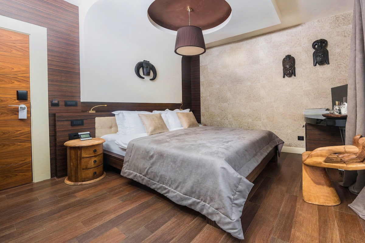 A modern bedroom with a large bed, wooden flooring, a round ceiling light, a small wooden side table, and a nightstand.