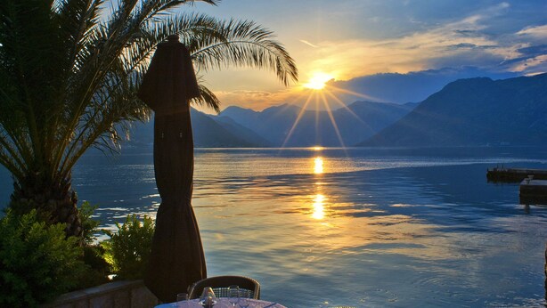 A sunset view over a calm lake with a dining table set for two, surrounded by wicker chairs and a palm tree. - Forza Mare (Kotor)
