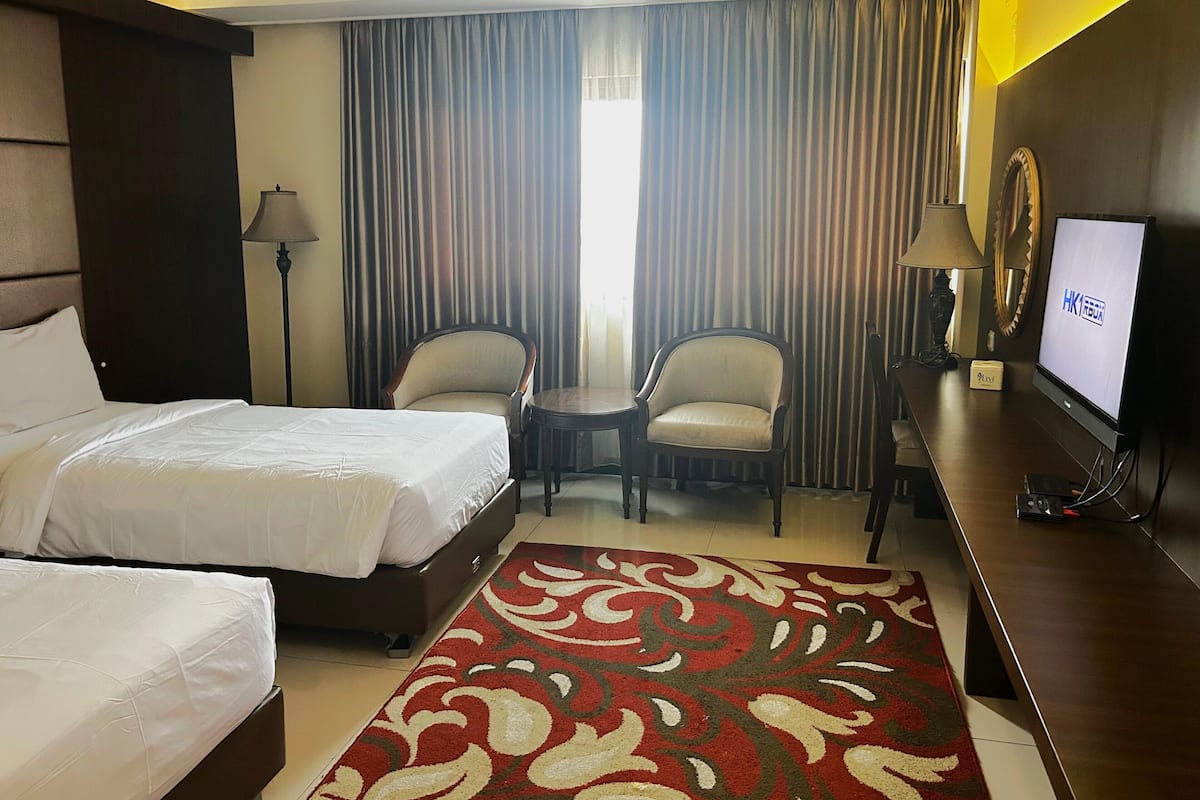 A hotel room with two beds, a desk with a television, a chair, and a large window with curtains.