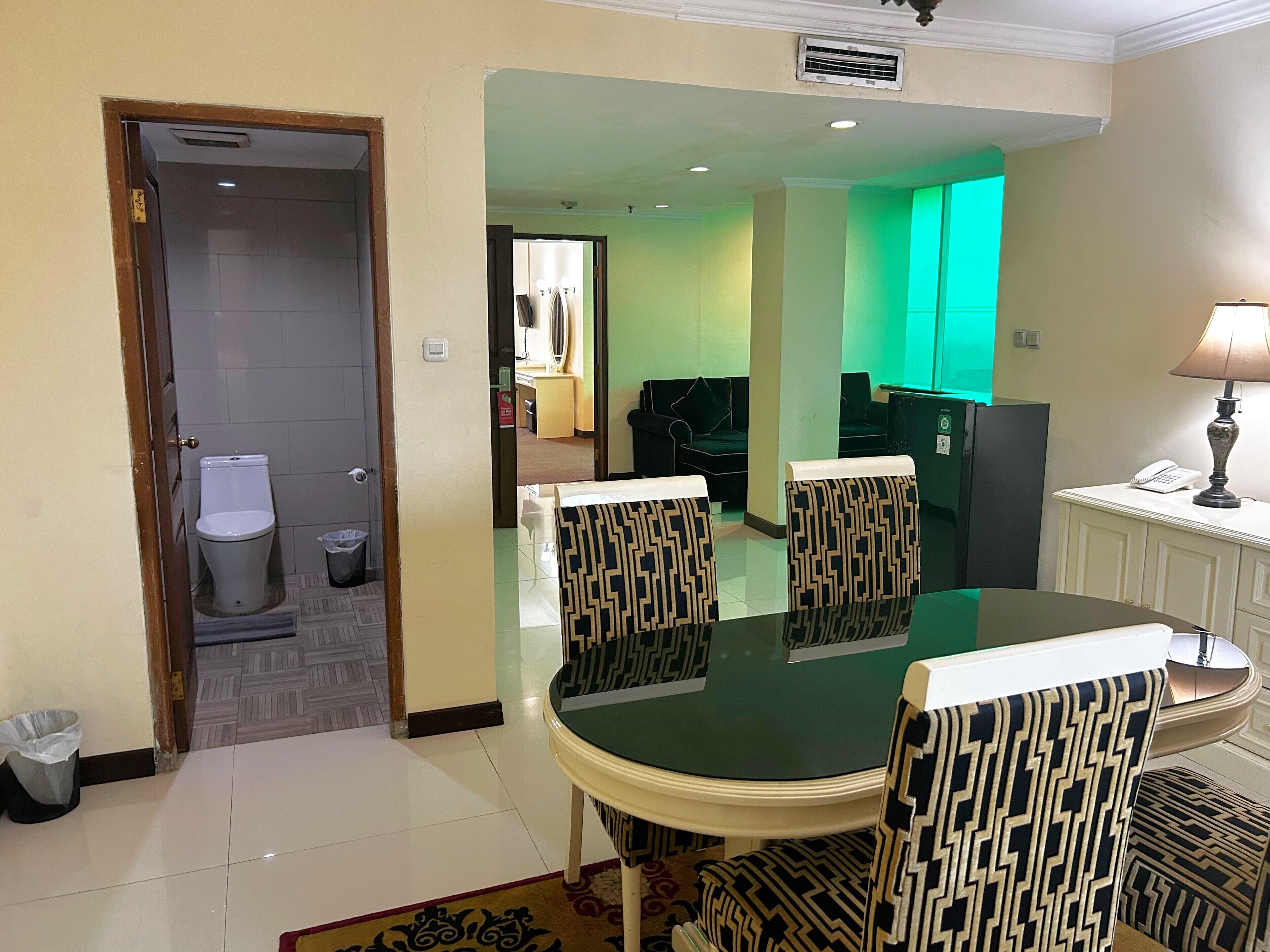 A hotel room with a dining table, chairs, a mirror, a sofa, a TV, and a bathroom visible through an open door.