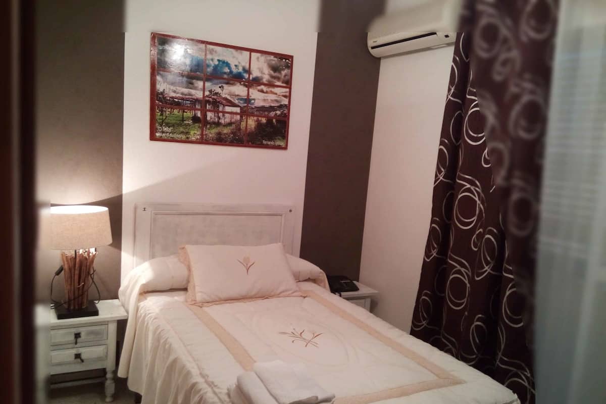 Un lit bien fait, recouvert d’une couvre-lit blanche et agrémenté d’oreillers, une lampe de chevet, une table de chevet avec une lampe, et un tableau accroché au mur.