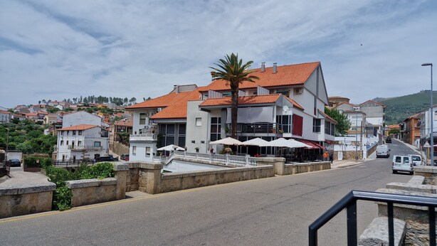 A street view with a building featuring a red-tiled roof, outdoor seating with umbrellas, and a stone wall. - Hotel La Palmera (Valverde del Fresno)