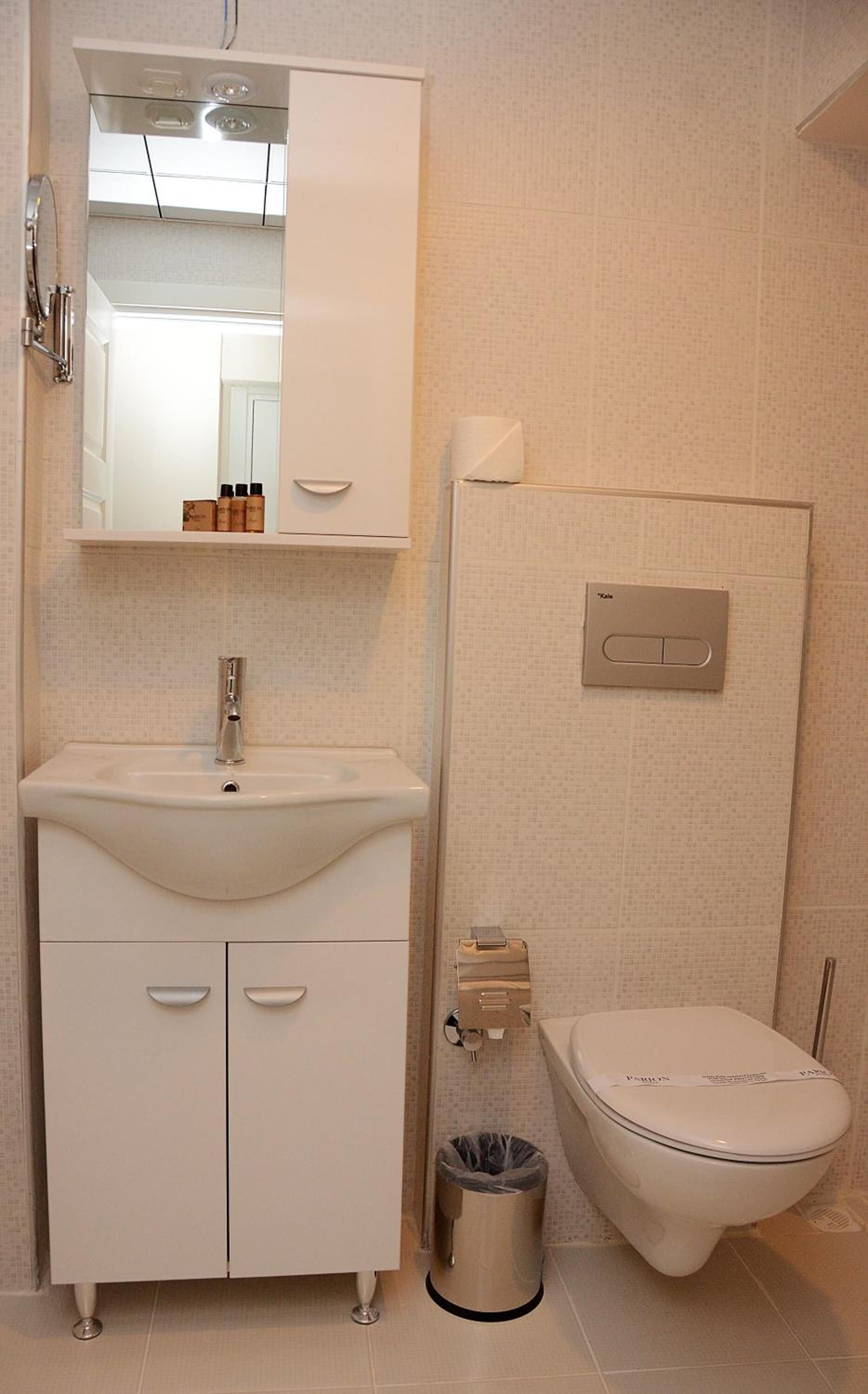 A bathroom with a white sink, mirror, and toilet. There is a trash can next to the toilet.