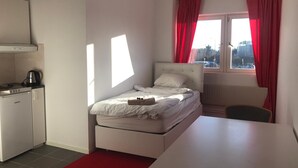 A compact hotel room with a bed, a small kitchenette, a desk, and a window with red curtains. - Patrician Hotel (Trangsund)