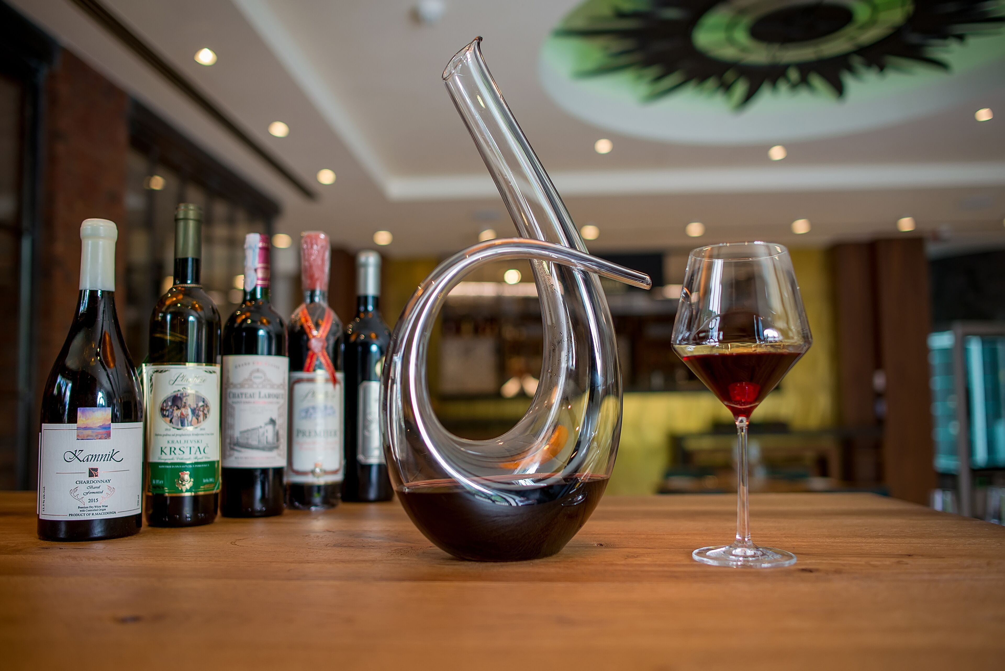 A wine bottle labeled 'Kammik' and a glass of red wine on a wooden table with other wine bottles in the background.