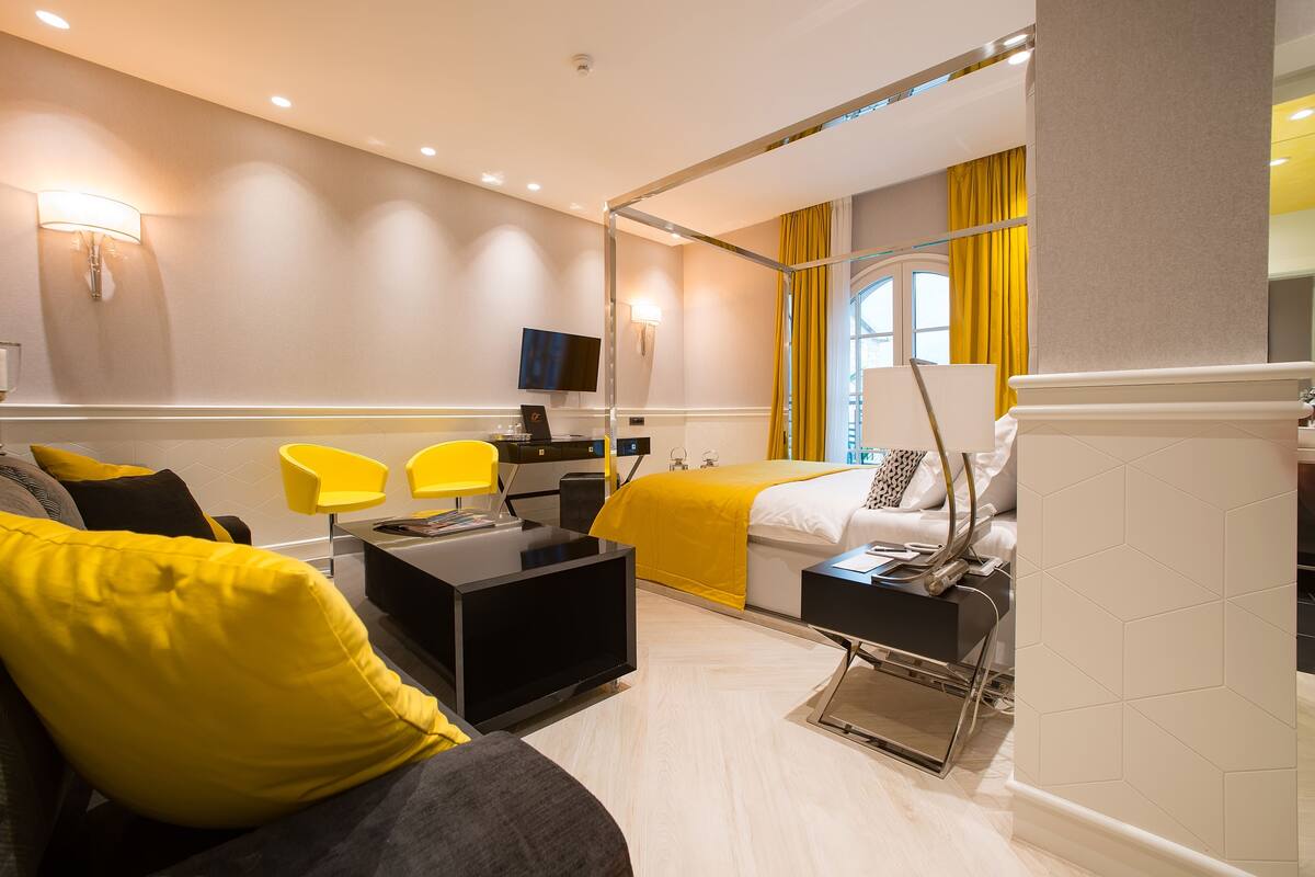A modern hotel room with a bed, a desk, a TV, and a seating area with yellow chairs.