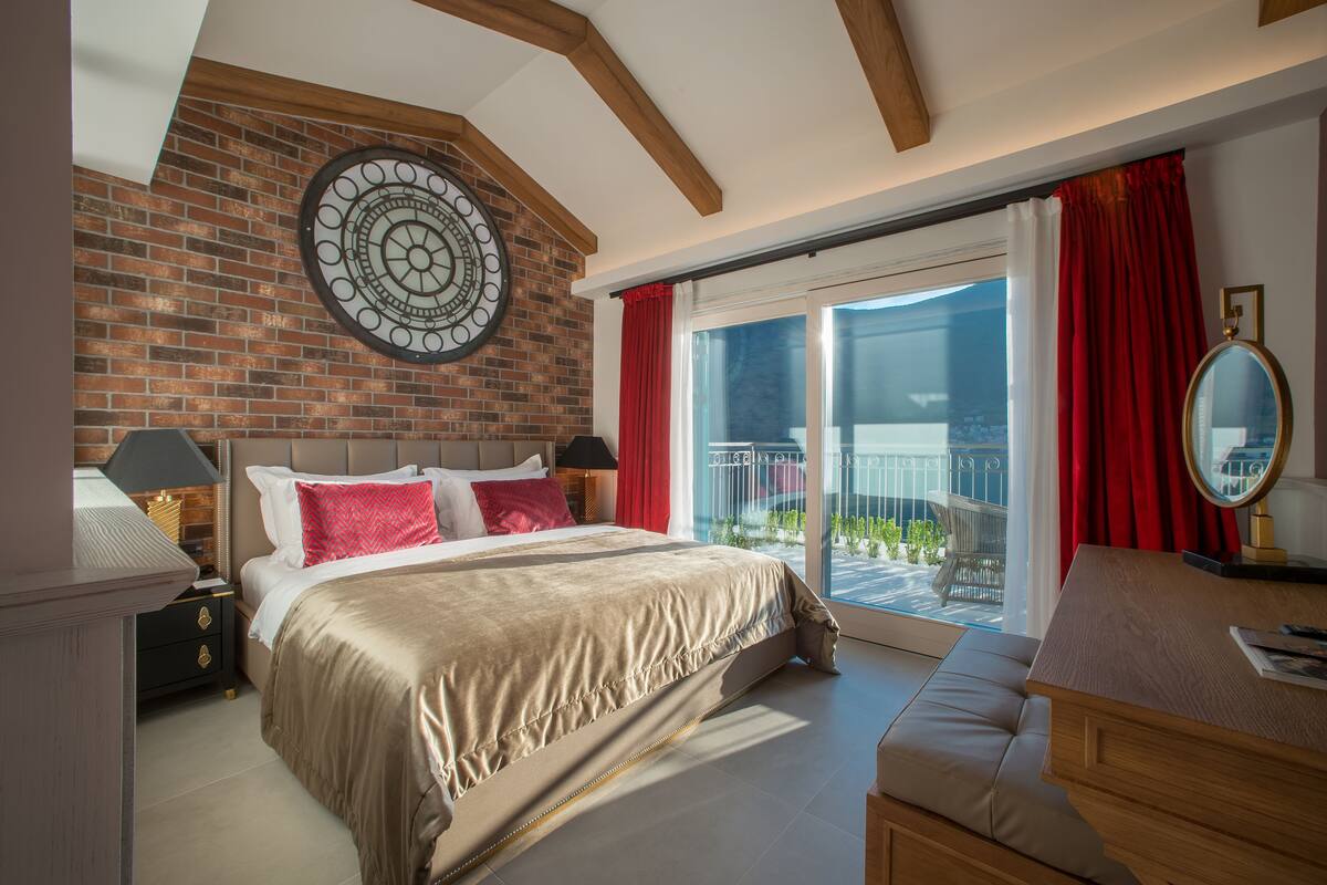 A bedroom with a large bed, a wooden headboard, a brick wall, a circular wall decoration, a balcony with red curtains, and a wooden dresser with a mirror.