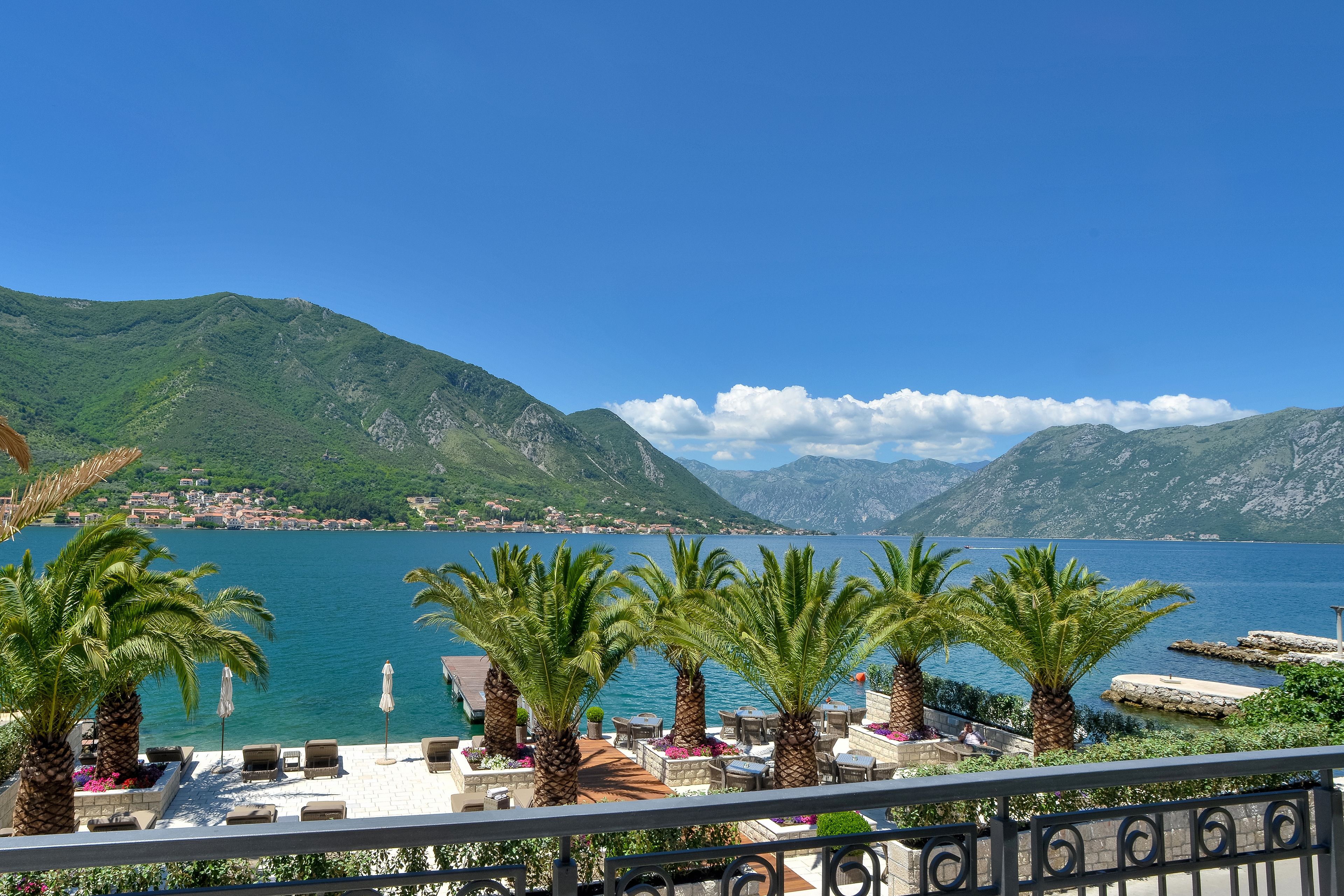 Una terraza con palmeras, mobiliario exterior y vistas a un lago y montañas.