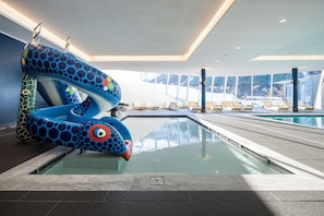 An indoor swimming pool with a colorful slide, lounge chairs, and large windows.