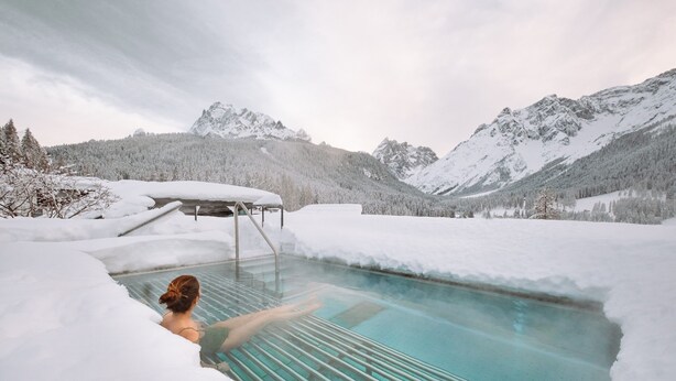 A person in a glass-bottomed pool surrounded by snow-covered mountains and trees. - Family Resort Rainer (Sesto)