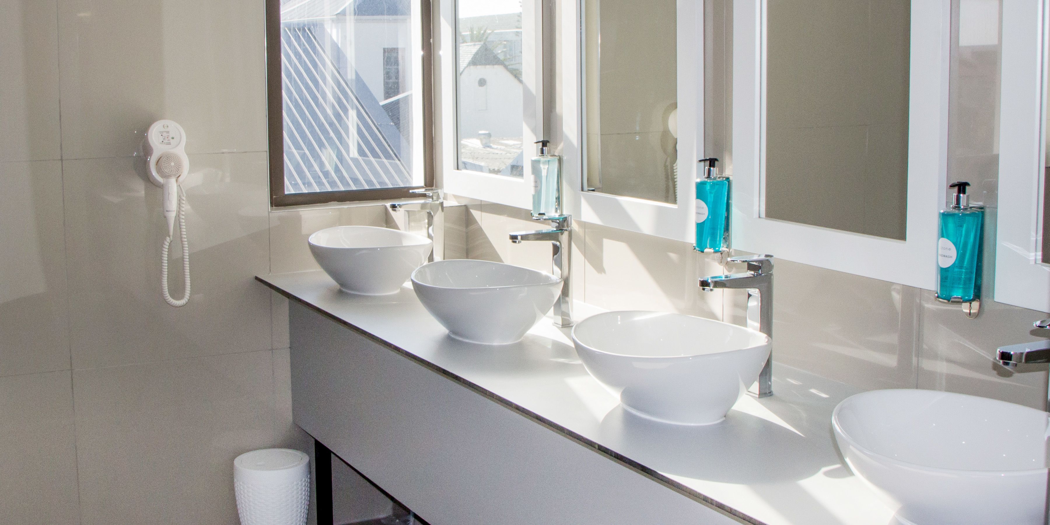 Ein Badezimmer mit drei weißen Waschbecken, einem Spiegel und einem Fenster.