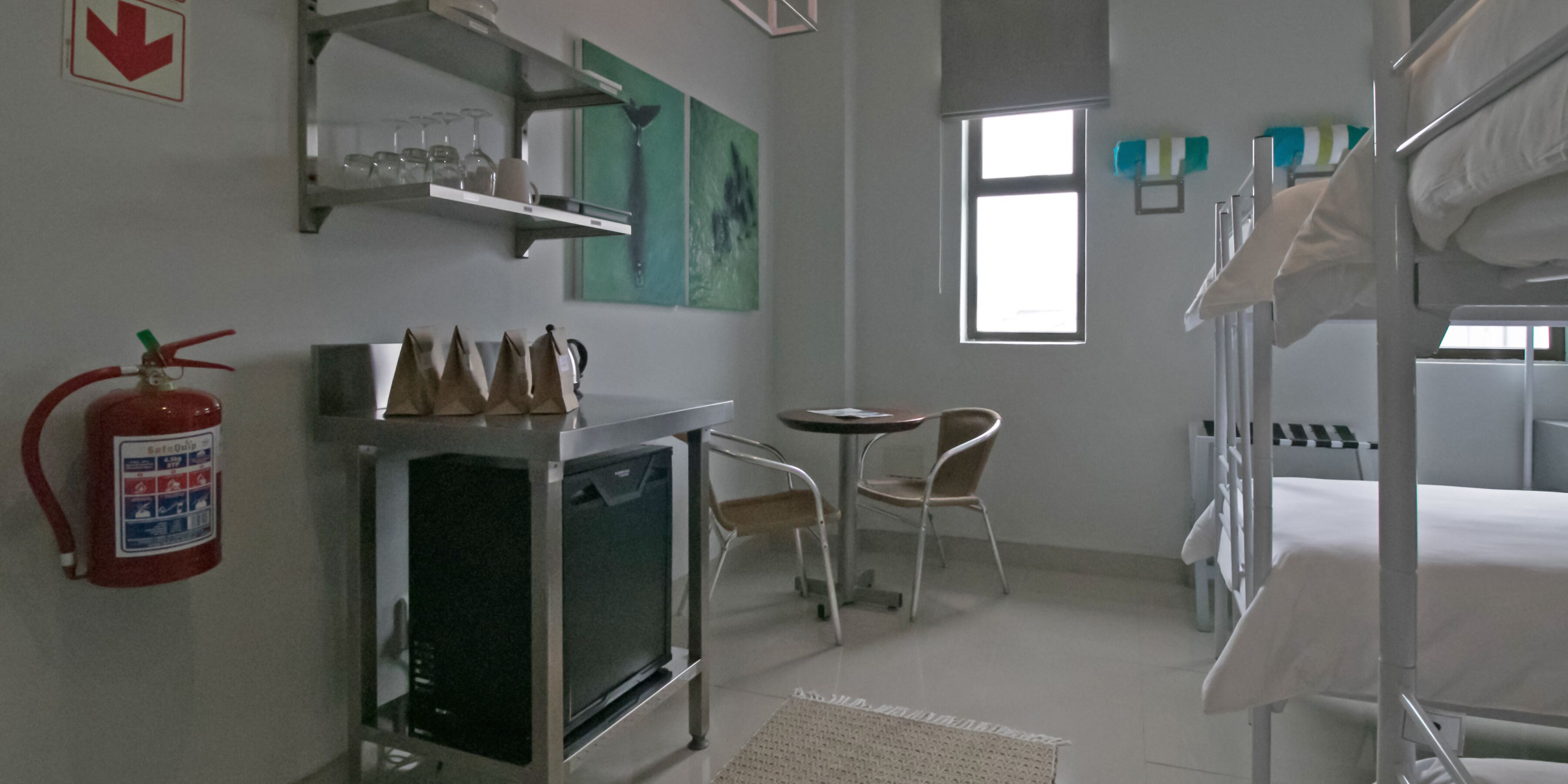 A hotel room with bunk beds, a small kitchenette, a table with chairs, a window, and artwork on the walls.