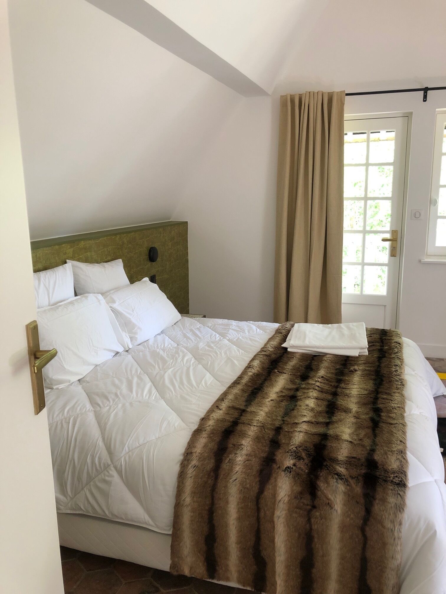 Un lit bien fait, avec du linge de lit blanc et un plaid en fausse fourrure marron, une tête de lit en bois et une porte ornée de rideaux beige.