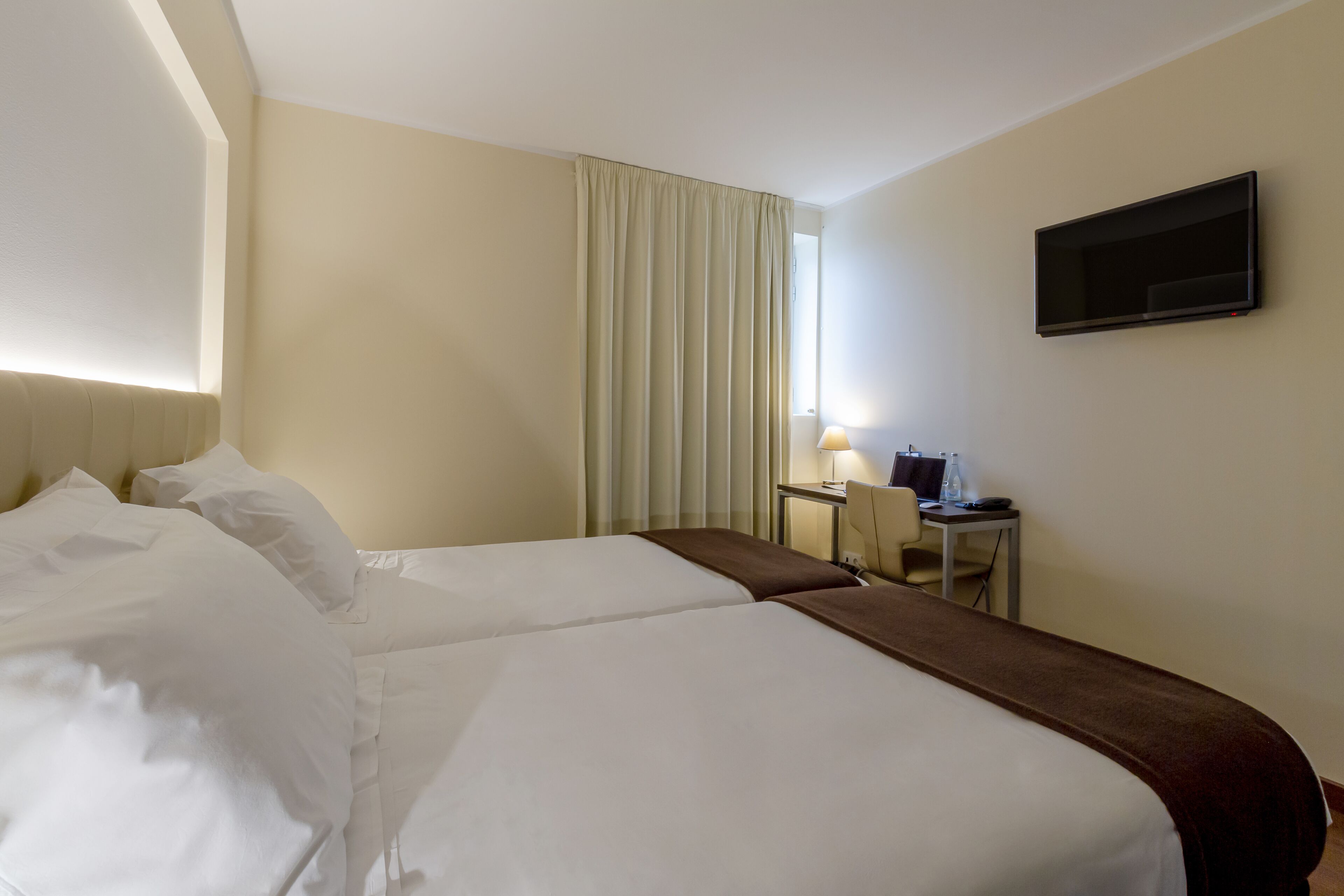 A hotel room with two beds, a desk, a chair, a TV, and a window with curtains.