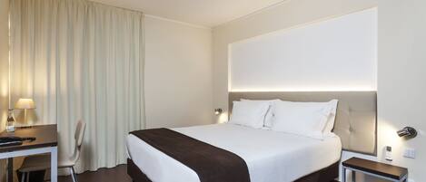 A hotel room with a large bed, a desk with a chair, a bedside table, and a lamp.