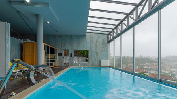 An indoor swimming pool with clear glass walls, a view of the city, and a modern interior design. - In Gold Hotel & Spa (Agueda)