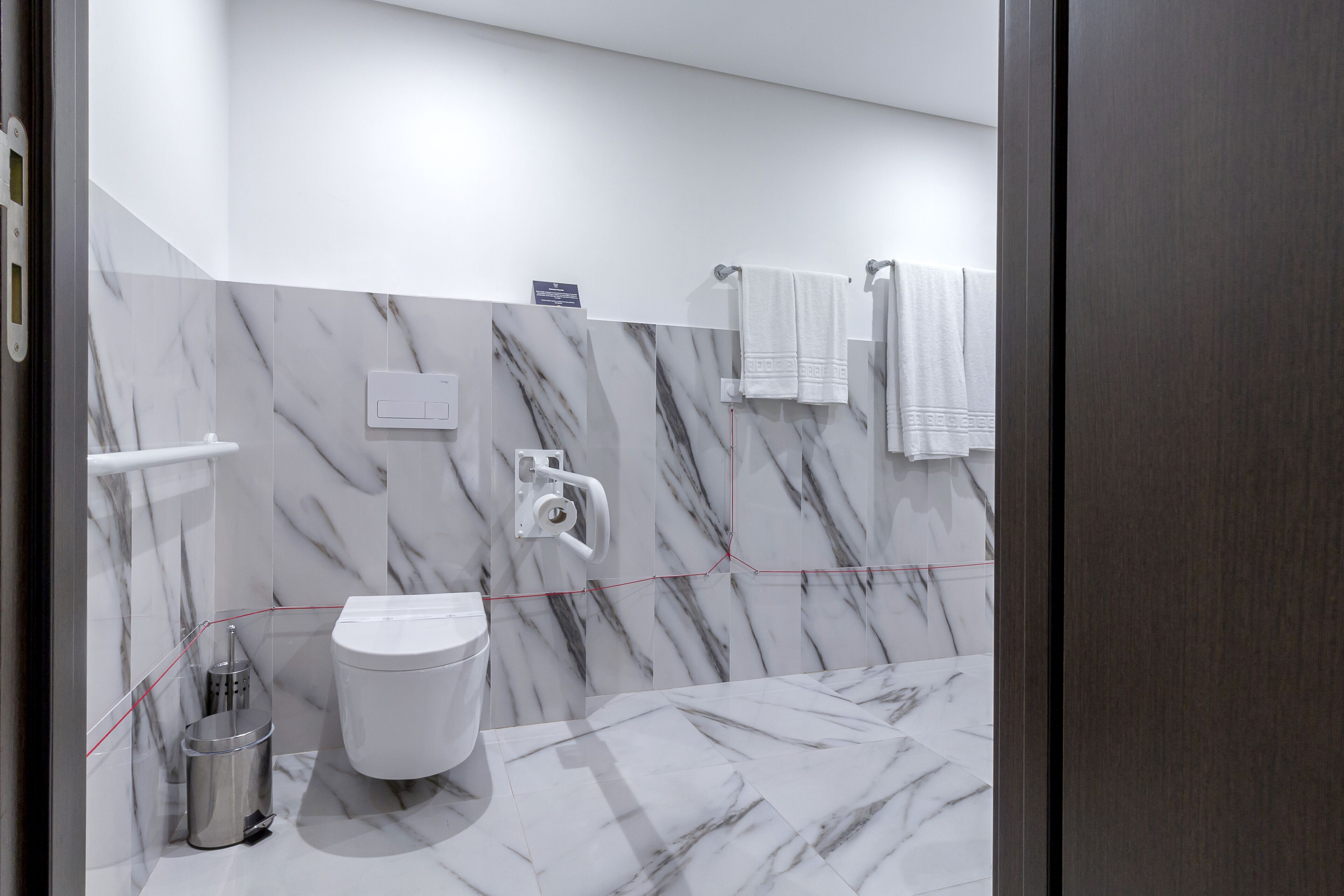 A modern bathroom with a marble floor and walls, a toilet, a hand dryer, and a trash bin.