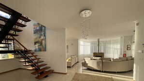 A modern living room with a staircase, a large abstract painting, a fireplace, and a large window with sheer curtains.