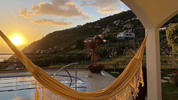 A hammock under an arched ceiling overlooking a pool and a scenic hillside view during sunset.