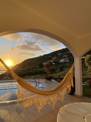 A hammock under an arched ceiling overlooking a pool and a scenic hillside view during sunset. - Luxury villa with private heated pool, garden and views of the sea and mountains (Arco da Calheta)