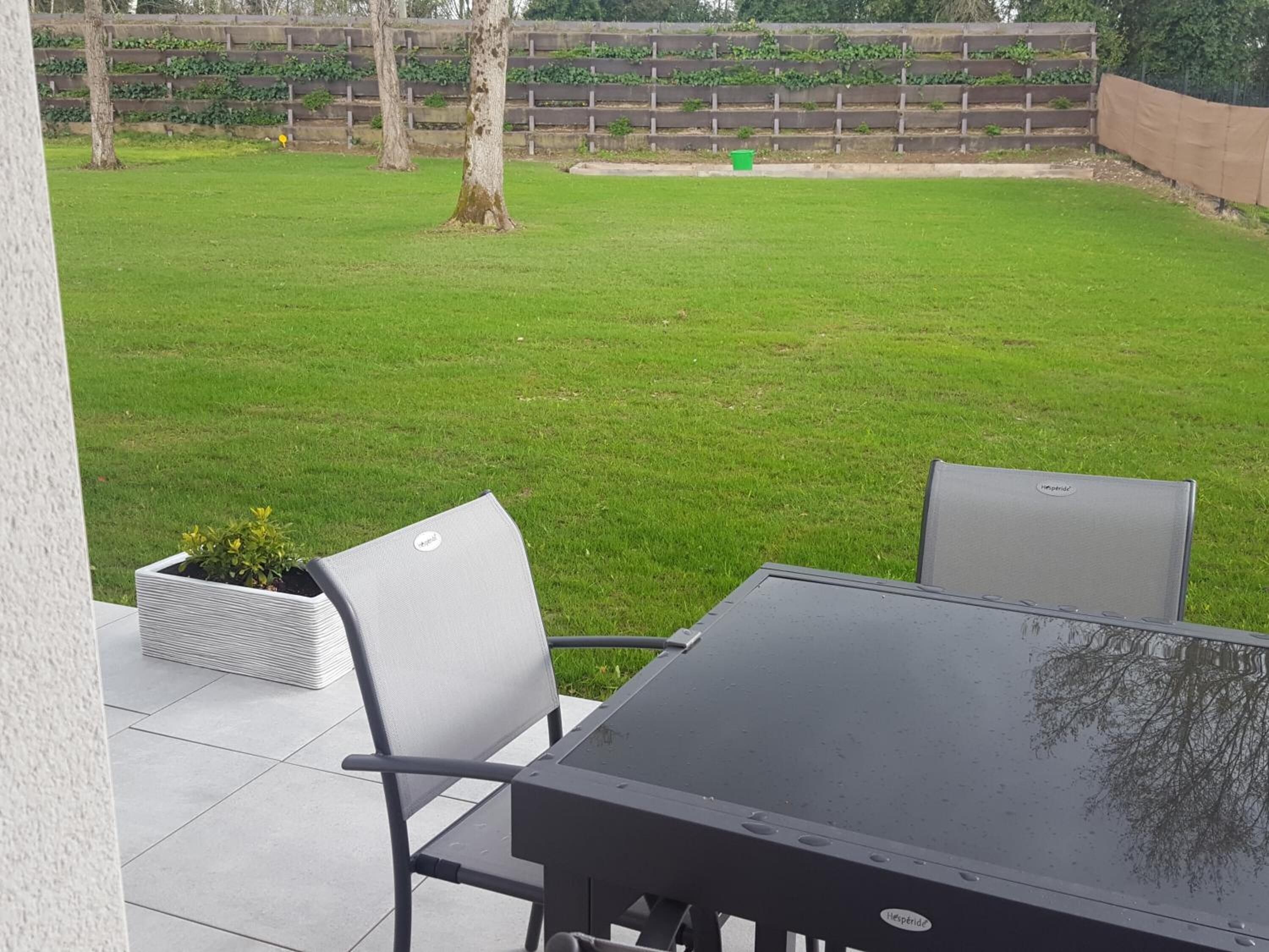 Un espace extérieur aménagé en patio, avec une table et des chaises, un petit jardinier avec des plantes, et une pelouse verdoyante bordée d’une clôture en bois et avec des arbres en arrière-plan.