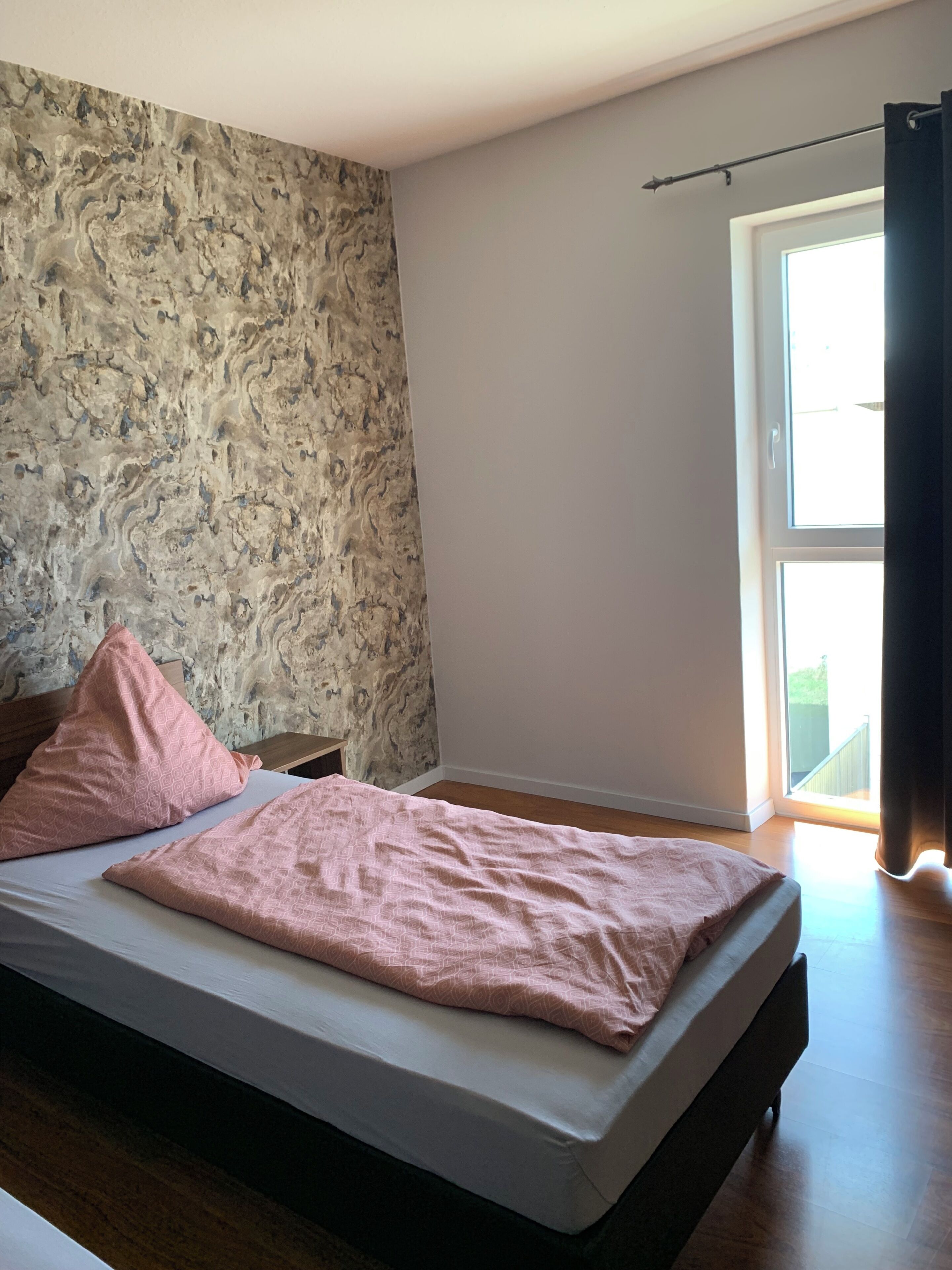 Ein Schlafzimmer mit einem Bett, einem Nachttisch, einem Fenster mit Vorhängen und einer Wand mit marmorartigem Muster.