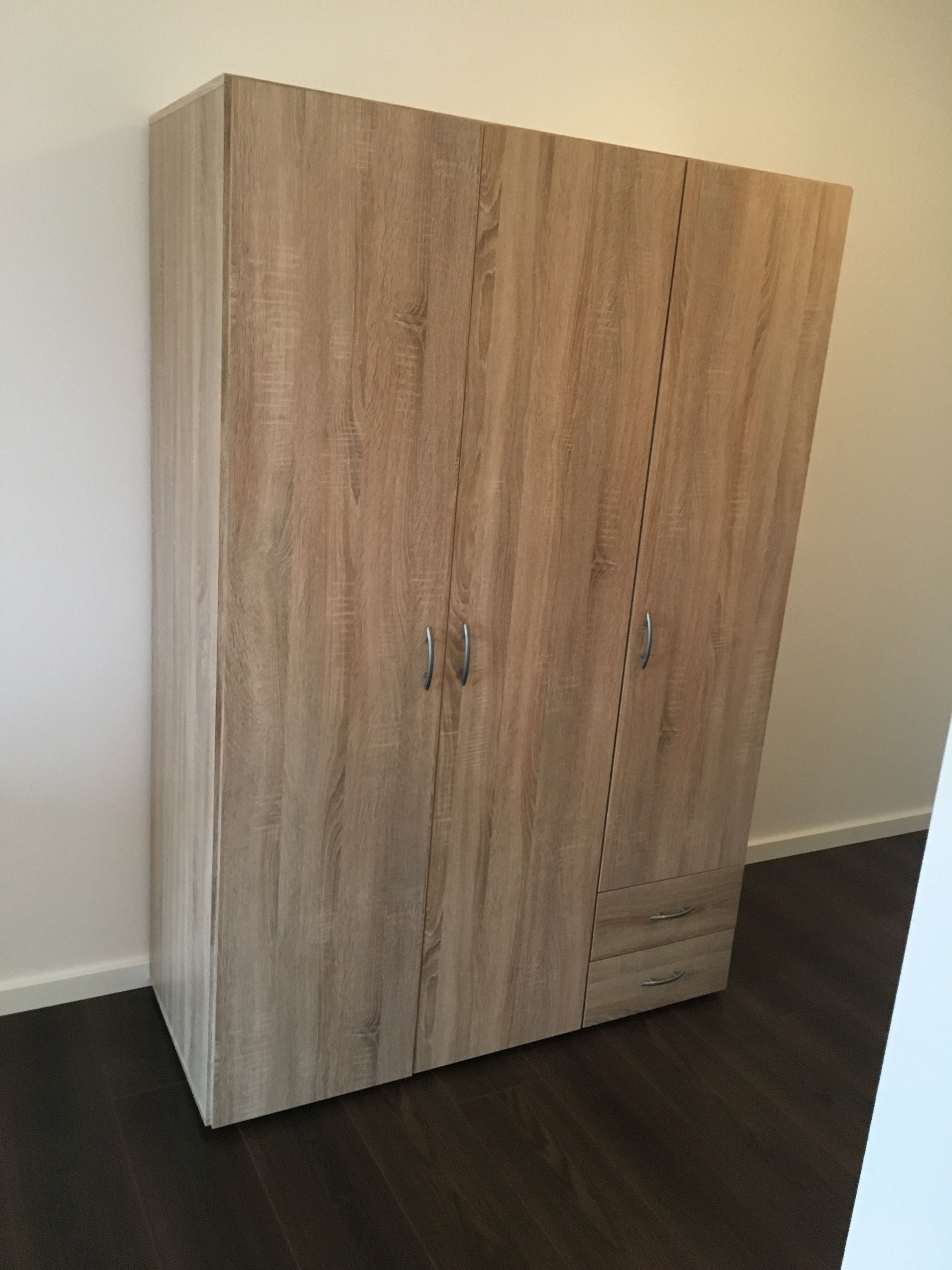 A wooden wardrobe with two doors and a drawer, positioned against a plain wall and dark flooring.