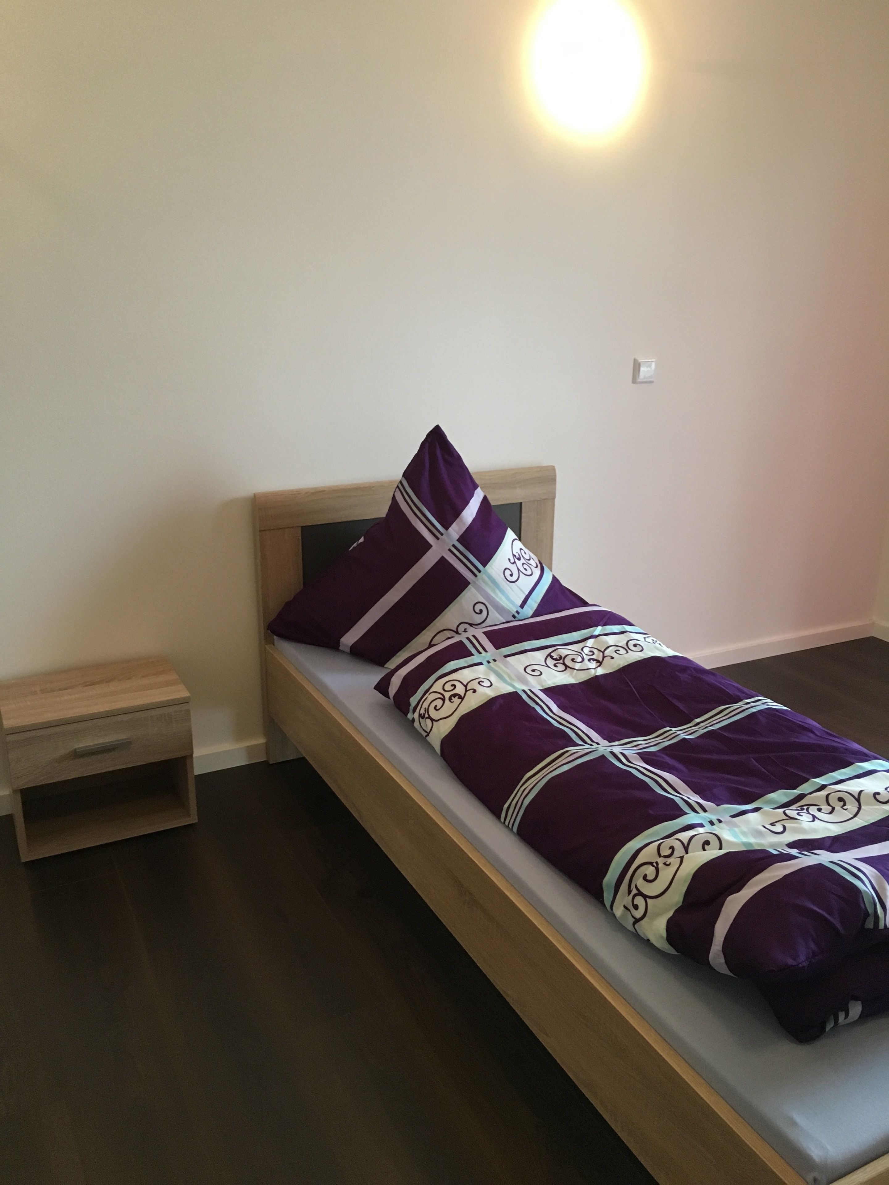 A single bed with a wooden frame, a nightstand, and a wall-mounted light.