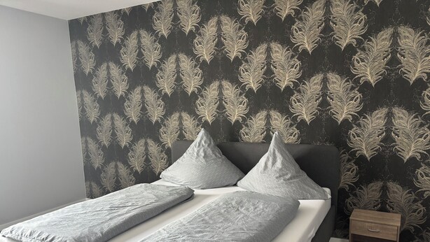 Een moderne slaapkamer met donker behang met patroon, een grijs bed met kussens met een dessin en een nachtkastje van hout.