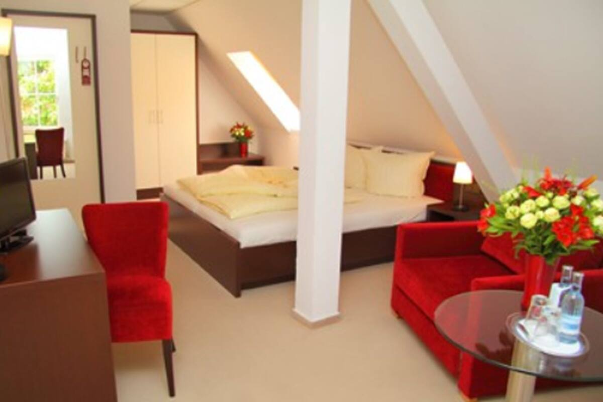 A hotel room with a bed, a desk with a chair, a red sofa, a glass coffee table, and a flower arrangement.