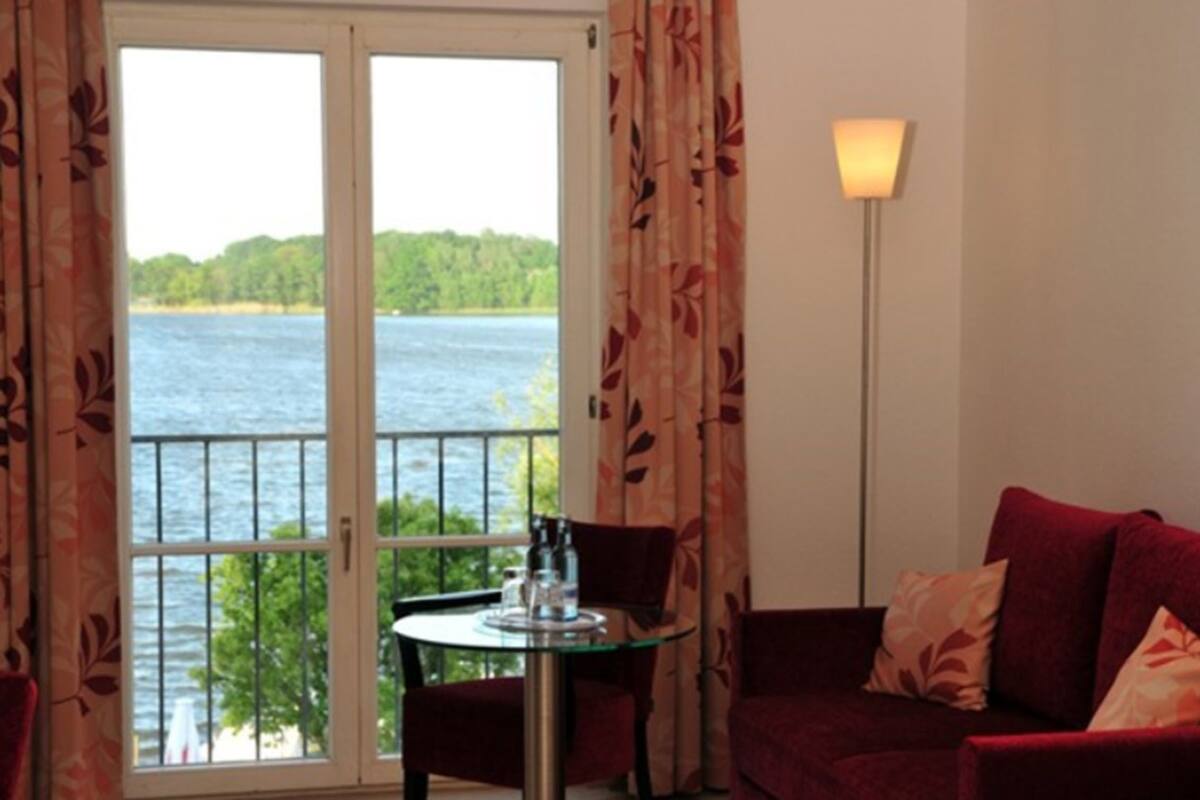 A room with a balcony overlooking a lake, a table with chairs, and a red sofa with patterned cushions.