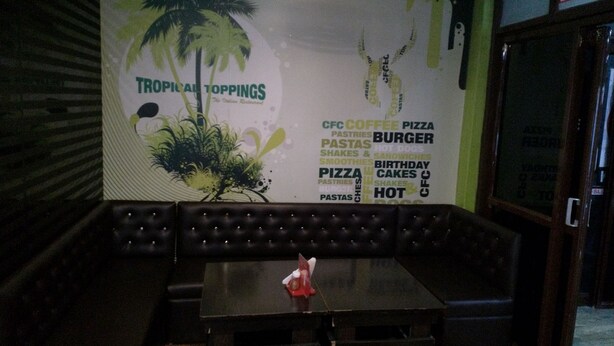 A restaurant interior with a booth seating area, a table with a red napkin holder, and a menu board advertising tropical toppings and various food items. - Tropical Suites (Phuntsholing)