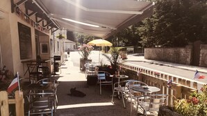 Ein Straßencafé mit Metallstühlen und -tischen, einem gelben Sonnenschirm und einer Steinwand.